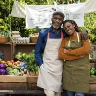 A Black man and woman stand together smiling in front of a fresh food stand to highlight the benefits of volunteering at a farm stand.