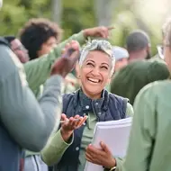 A woman with short, silver hair smiles at a group of volunteers she is managing as part of a corporate volunteer effort. They're all wearing dark green long-sleeve shirts and standing outside, with trees in the distance.