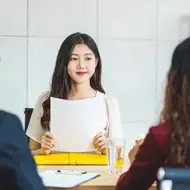 A young woman of Asian descent sits at a long table in front of two hiring managers—a man with short hair and a woman with long curly hair—holding a piece of paper in front of her.