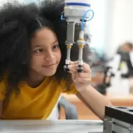 A young Black girl sits at a desk in a school, looking at a instrument in science class. Behind her are microscopes and other scientific instruments.