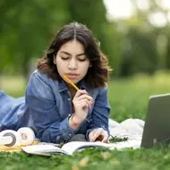 A photograph of a young woman sitting on grass while studying with open books and a laptop.