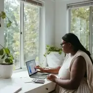 Imagem de uma mulher afrodescendente de branco e faz contraste com a sala que é toda branca. Ela está sentada na frente de um notebook com uma calculadora, alguns papéis. Está em uma mesa próximo da janela, é uma sala bem iluminada e cheia de plantas que podemos ver através da janela e dentro da sala em vasos.