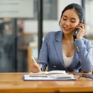 Woman talks on phone in office while writing in notebook.