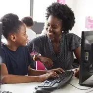 A Black mother and daughter smile at each other while sitting by a computer.