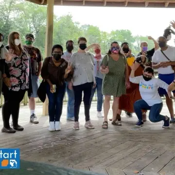 A group of multiracial people outdoors in a gazebo wearing face masks. Some are waving and some are making silly faces or gestures/poses. Fresh Start's logo is in the bottom left corner.