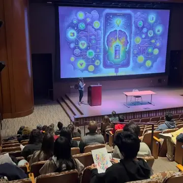 A speaker presents in a lecture hall with a large brain-themed infographic projected behind them.