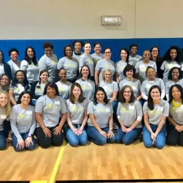 A large group of women in a school gym all wearing a gray t-shirt with a logo on the front. They are lined up in three rows, standing, sitting, and then kneeling for a group photo.