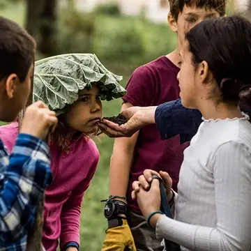 Child with large leaf on head smells a handful of soil held by teacher as classmates observe