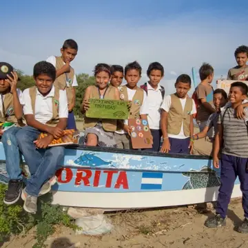 A group of children in uniform pose on a boat at the beach with a sign advocating to save sea turtles.