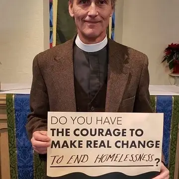 A clergy person holding a sign that reads "Do you have the courage to make real change to end homelessness?"