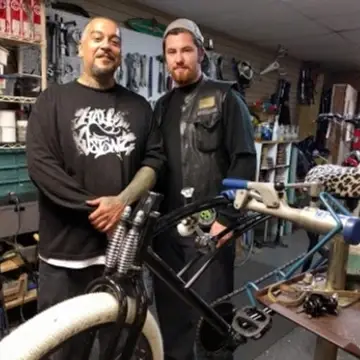 Two men pose with a chopper bicycle they are fixing.