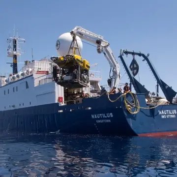 Exploration Vessel Nautilus viewed from the port quarter painted blue near the waterline and white above decks is launching a yellow Remotely Operated Vehicle using a white crane