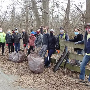 A group of people wearing masks stand with trash bags and litter grabbers next to a fence. Their eyes are smiling.