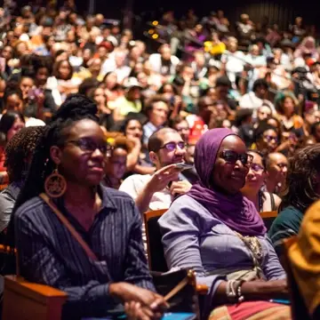 A large audience looks at something to the right, off camera. In the foreground, in focus, are two Black people — one older than the other and wearing a headscarf.