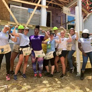Ten women standing in front of a house being constructed making a muscle with one arm in a group photo. Some women are wearing hardhats and all are dressed in t-shirts and leggings, jeans or shorts.