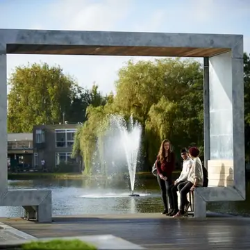 Students chatting on metal sculpture/bench in front of a lake with a fountain in the background