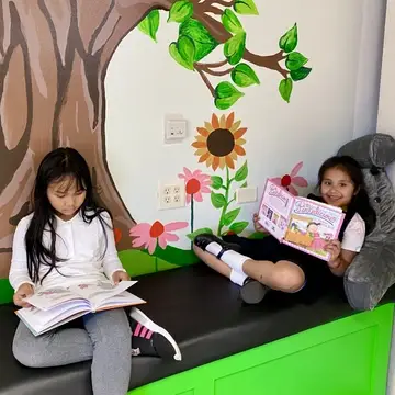 Two young girls reading books on a new bench under a mural of a tree and flowers.