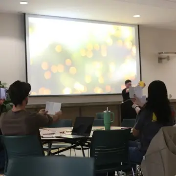Students in a university setting raise papers to critique a presentation with soft bokeh lights projected in the background.