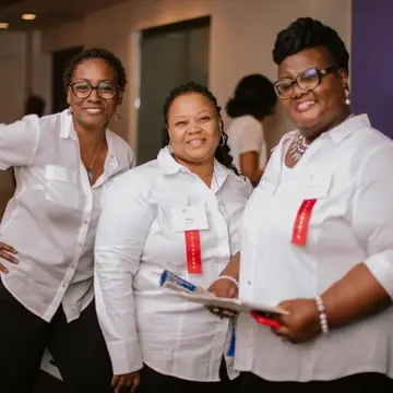 Three smiling Black women in white blouses and black pants with nametags that have a red ribbon that says volunteer at a social event.