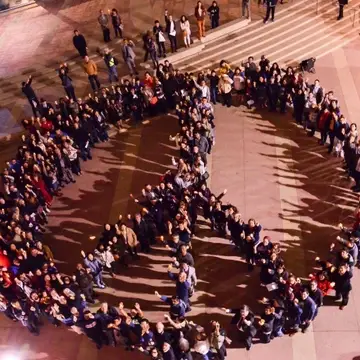 picture of a group of people creating a peace sign in a courtyard while holding their hands up. The picture is on a brown floored courtyard and there are about 300 people in the formation. The photo was taken from a bird's eye view.