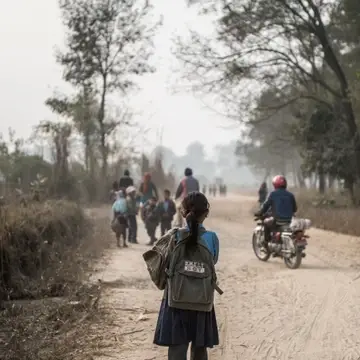 Young girl walks down busy dirt road carrying two backpacks
