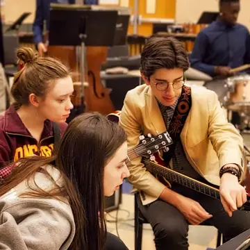 A young man holding a guitar points his finger at a music stand while two young women, also with guitars, look on.
