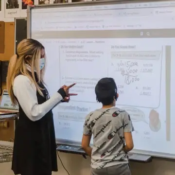 A teacher and a student stand by a SMARTBoard for a math lesson. The student stares at the board and the teachers signs.