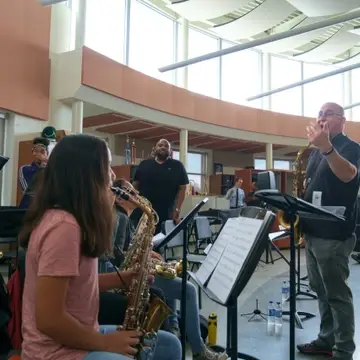 A man with a saxophone gesticulates in front of seated students playing saxophones.