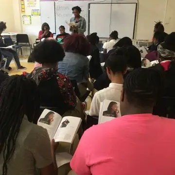 a teen author reads her book to a class full of students. The students have copies of the. book and are looking at the page.