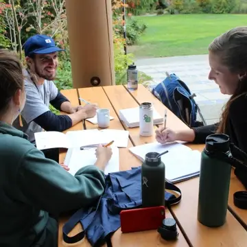 Three smiling graduate students sit at an outdoor table doing homework
