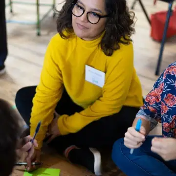 A young woman  in a yellow sweater at a facilitation and collaboration listens poised to write on a post-it.