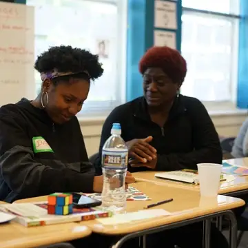 Two Black women talking. One women is writing while the other uses her hands to make a point.