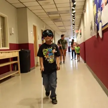 A child walks down a hallway wearing learning shades and using a white cane. Other adults and children are in the background with canes in hand.