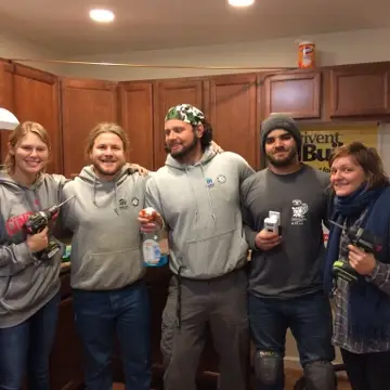 Five young adults posing for a photo with a drill, cleaning products, and coffee cups
