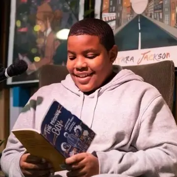 kid in light colored hoodie on the mic grinning and reading from a book
