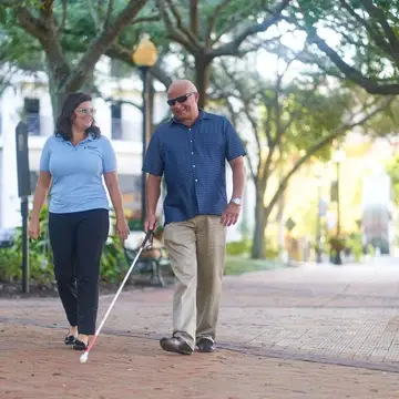 A man walking with a white cane and a woman walking beside him smiling. In a park.