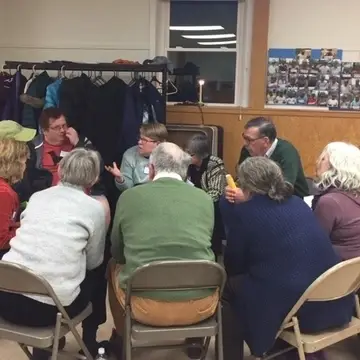 A group of 12 adults gather in a circle on folding metal chairs in the basement of a church to talk.