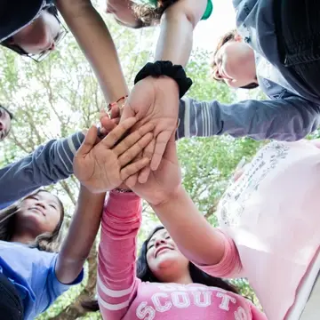 7 Girl Scouts stand outside in a circle with their hands coming together in the center of the circle.