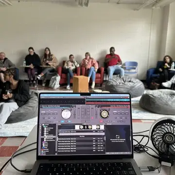 A view from behind a laptop showing a DJ software interface, with a group of students seated casually around a classroom.