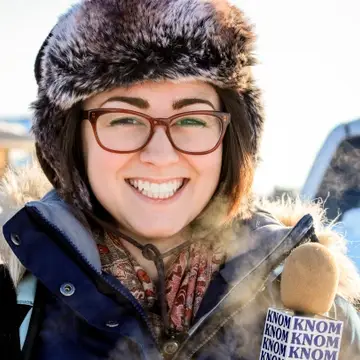 Woman in heavy parka smiles while holding "KNOM" microphone