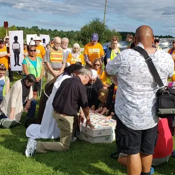 Clergy kneel as they pray over a mock casket with pictures of undocumented people they know are attached to the outside. They are surrounded by others in orange LA RED shirts with media present.