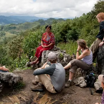 Guatemalan woman sits on a rock overlooking a valley, talking to 4 western students who are sat listening