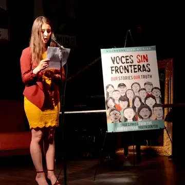 a teen author stands on stage reading into a microphone. The cover of her book "Voces Sin Fronteras" is next to her.