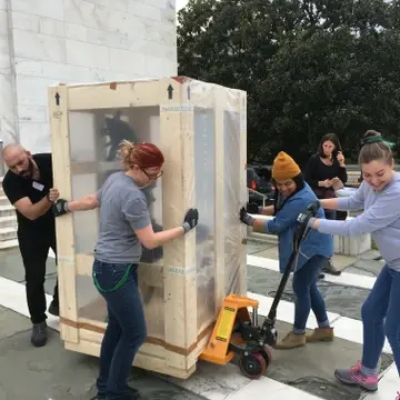 5 Folger staff working together to bring out exhibits; hauling a large exhibit object outside in front of Folger steps prepping for renovation