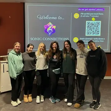 A group of seven smiling young adults stand in front of a presentation screen welcoming attendees to "SONIC CONNECTIONS" with a QR code for a pre-event form.