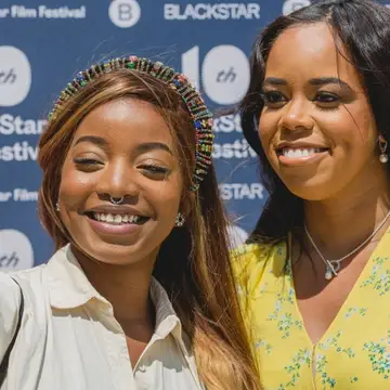 Two Black people pose for a selfie at the 10th BlackStar Film Festival, in front of a blue step-and-repeat banner that displays the festival name.