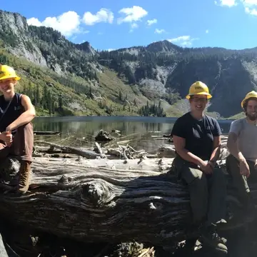 Two groups of three people sit side by side on a large log near an alpine lake; five are wearing yellow hard hats and one is wearing a white hard hat.