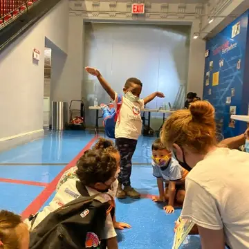 5 year old dark skinned boy wearing a Dance Project t-shirt acts out the movements of the book being read by Executive Director. Other small children are sitting nearby listening to book