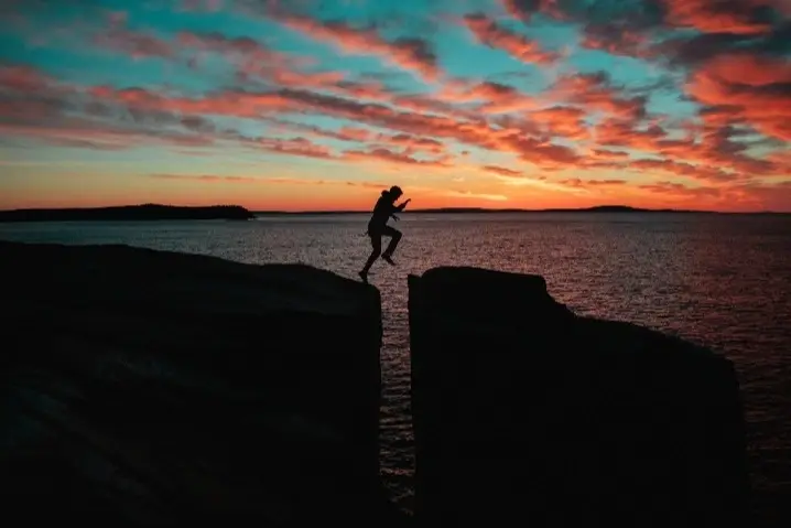 Al fondo un atardecer en el mar y una persona saltando de una roca alta a otra