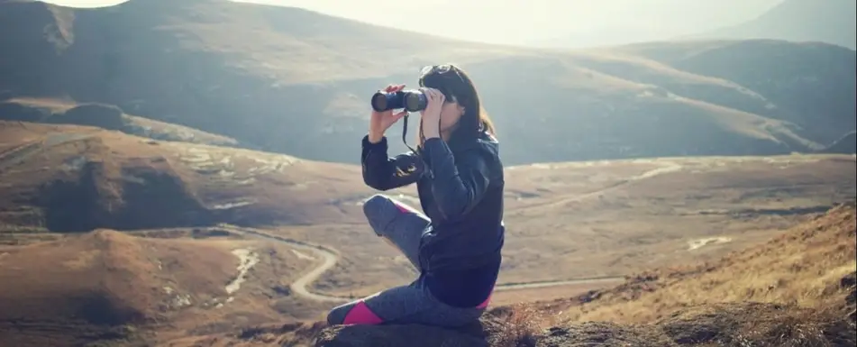 A person sitting on a cliff using binoculars.
